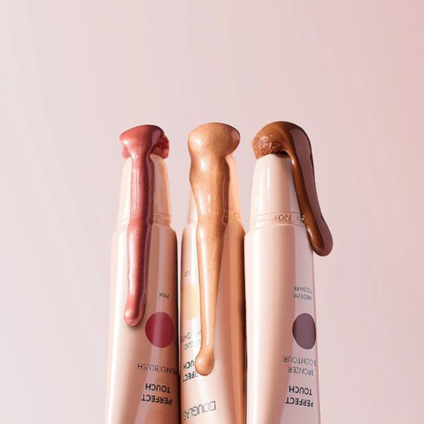 Kolm DUOGAS Perfect Touch kreemjat meigitoodet – põsepuna, highlighter ja kontuurimisvahend – pastelroosal taustal, toodete peal säravad tekstuurinäidised.