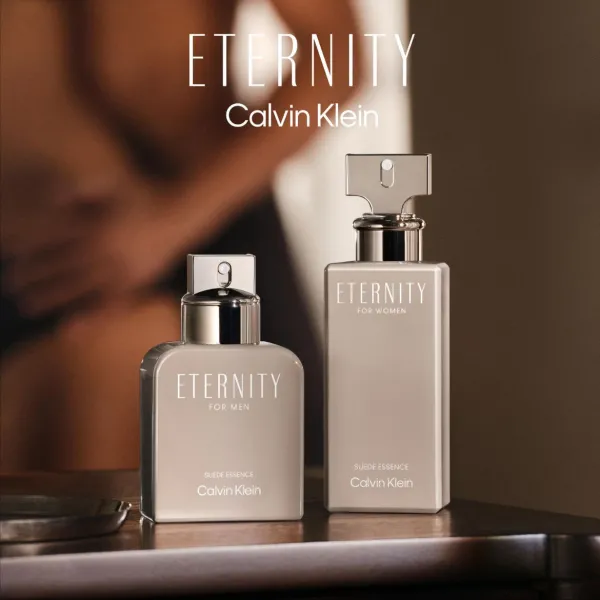 Kaks Calvin Klein Eternity Suede Essence lõhnapudelit, üks meestele ja teine naistele, on paigutatud lauale soojades pruunides toonides interjööris. Esiplaanil on näha metallsete detailidega tooted, taustal aga hägustatud interjöörielemendid ja inimese siluett.