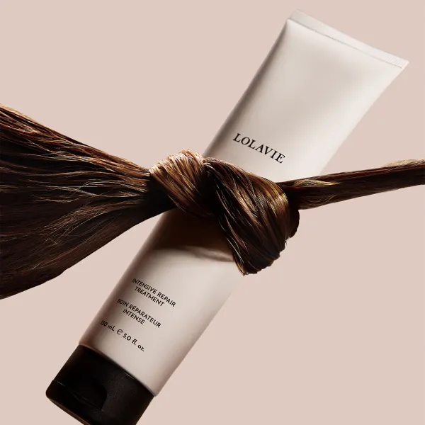 LolaVie Intensive Repair Treatment juuksehooldustoote tuub on kujutatud heledal beežil taustal. Toodet ümbritseb läikivast juuksesalgust seotud sõlm, mis rõhutab visuaalselt toote taastavat ja tugevdavat toimet juustele.